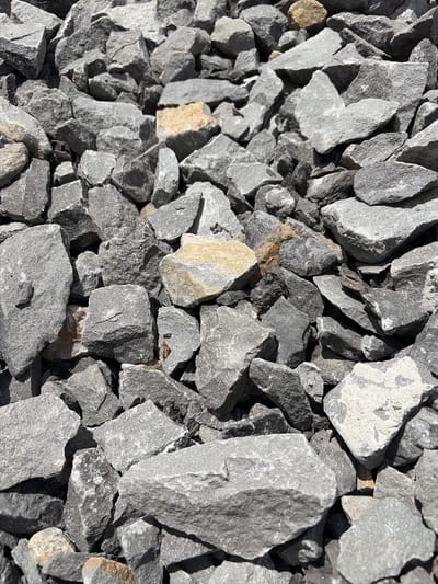 This is a close-up, overhead, static shot of a pile of grey, rough-edged gravel. The gravel consists of angular, irregularly shaped rocks of varying sizes, with a few lighter-colored, tan rocks interspersed within the predominantly grey mass. The lighting suggests a bright, possibly sunny environment, casting shadows that emphasize the texture and three-dimensionality of the stones. The scene is devoid of any human presence, activity, or discernible urban or natural environment beyond the gravel itself. The image does not depict Janowice, Poland or any identifiable geographical location.