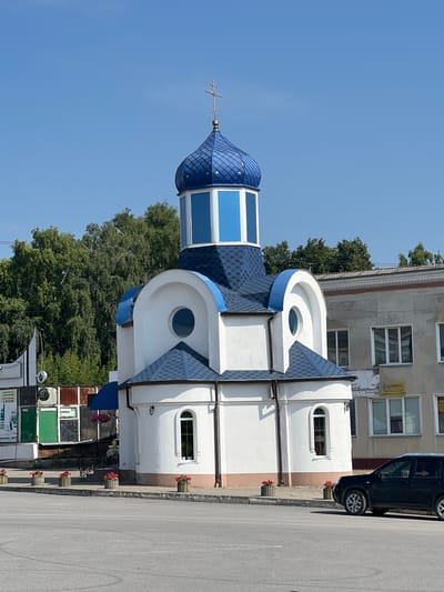 The image shows a small white church with a blue domed roof and a golden cross on top, located in the city of Chemerivtsi, Ukraine. The church features arched windows and is surrounded by a paved area with flower planters. To the right of the church, a multi-story building with windows and signs is visible. A black SUV is parked in the foreground on the right side of the image. In the background, trees and a clear blue sky can be seen. The overall scene depicts a sunny day in what appears to be a town or city setting.