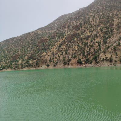 A wide-angle, eye-level shot captures a scenic view of a body of water in front of a steep, rocky mountain slope. The water in the foreground is a vibrant green, with subtle ripples across its surface indicating a gentle breeze. The mountain slope, dominating the upper two-thirds of the image, is characterized by its arid, rocky terrain, with sparse vegetation in the form of scattered trees and shrubs. The colors of the mountain range are earthy tones of brown, tan, and rust, with patches of darker green from the vegetation. The sky above is a pale, hazy blue, suggesting a slightly overcast or dusty atmosphere. The location is identified as Tabaroucht, Morocco. There are no people or discernible man-made structures in the frame, making it a purely natural landscape.