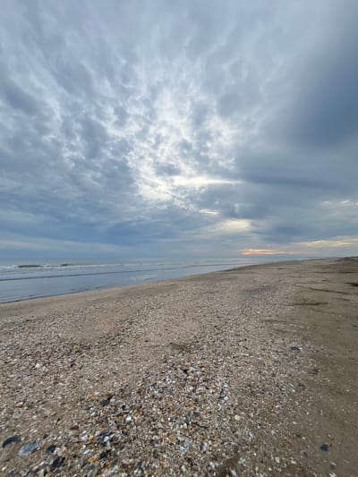 The image displays an expansive, uninhabited beach and ocean scene under an overcast sky, located in the city of Alueduamuka O, Indonesia. The upper two-thirds of the frame are dominated by a dense, grey cloud cover, consistent with stratocumulus formations, with brighter, diffused light emanating from obscured areas in the central and right portions, suggesting the sun's position. A subtle, light orange-yellow tint is visible along the distant horizon on the far right, indicating either dawn or dusk conditions.

The horizon line is clearly defined, separating the sky from the body of water and the coastline. The ocean, occupying the left mid-ground, exhibits calm, grey-blue water with minor waves breaking gently onto the shore. A slender, dark, vertical object, possibly a distant marker or buoy, is faintly discernible on the water's surface in the mid-left distance.

The foreground and mid-ground of the beach consist of a wide, light-brown sandy expanse heavily mixed with a multitude of small, multi-colored shells, pebbles, and shell fragments, creating a rugged and varied texture. This shell-laden strip transitions to a broader area of darker, finer sand on the far right, which appears largely free of debris. There are no individuals, animals, structures, or any discernible human activity present within the depicted scene.