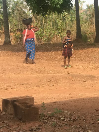 Two women stand on a dirt path in a rural setting. One woman balances a bucket on her head, wearing a red shirt and blue patterned wrap. The other woman carries a baby in her arms and wears a plaid skirt. Trees and vegetation surround them. A small brick structure sits beside the path. The ground is brown and dusty.