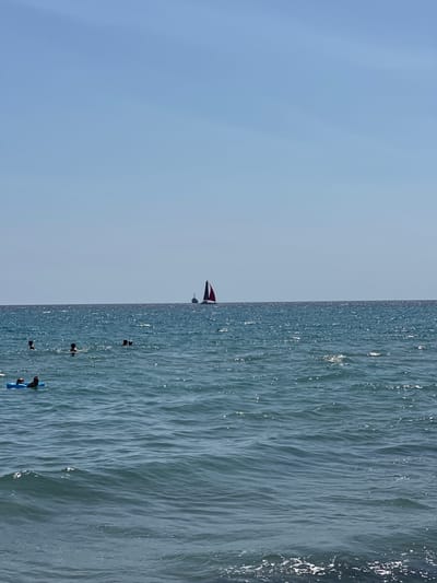 The image depicts a body of water, likely the sea, under a clear blue sky. In the foreground, several individuals are visible in the water. One person is floating on a blue inflatable raft, appearing relaxed with their legs propped up. Other figures are swimming or wading in the water at various distances. In the middle ground, a sailboat with a red sail is visible on the horizon, accompanied by a smaller vessel. The water is choppy with small waves, and sunlight reflects off its surface, creating a shimmering effect. The overall scene suggests a recreational activity taking place at a beach or coastal area. There is no discernible indication of the city of Biddeputzi/Villaputzu, Italy within the image.