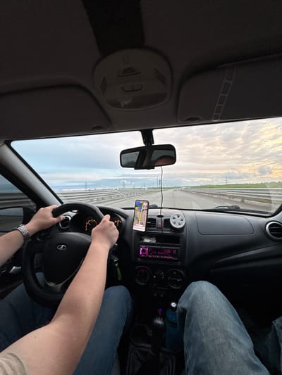 The image captures the interior of a car moving on a highway, likely taken by a passenger. The driver's hands are visible on the steering wheel, wearing a watch on their left wrist. A smartphone is mounted on the dashboard, displaying a map application. The dashboard features a radio and various controls. Outside the car, the landscape shows a highway with parallel lines and structures resembling electrical pylons under a sky with scattered clouds. Considering the query, the location is approximated to be Bol'shaya Mos', Russia, but this is inferred from the user's query and cannot be definitively confirmed from the image alone. There are no interactions visible between people; the primary action is driving. The overall environment is a car interior and a highway landscape.
