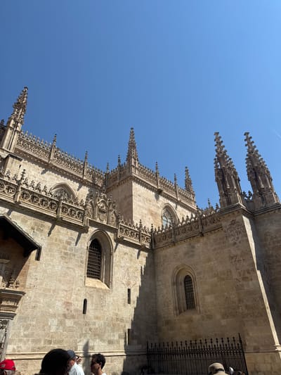 This is a low-angle outdoor, eye-level shot of the Granada Cathedral's facade in Granada, Spain, under a clear blue sky. The building is constructed of light-colored stone blocks and features Gothic architectural elements, including intricate carvings along the roofline, pointed spires, and arched windows with iron grates. Some of the stone walls show signs of wear and what appears to be graffiti or inscriptions. In the foreground, the backs and sides of several people are visible, looking up towards the cathedral. They are dressed casually, suggesting a tourist or visitor experience. The sunlight casts strong shadows across the building and the ground. The overall impression is of a historic and grand religious structure on a bright, sunny day.