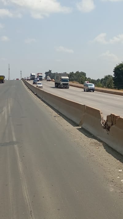 The image depicts a multi-lane highway in Sigona ward, Kenya, under a hazy sky with scattered white clouds. The perspective is from the left carriageway, looking forward and slightly right across a median. The immediate foreground displays an asphalt road surface with minimal debris and faint tire marks. To the right, a dirt and gravel shoulder borders a line of pre-cast concrete barriers. One barrier segment on the far right exhibits significant impact damage, exposing fractured concrete and rebar.
Traffic is active on both visible carriageways. On the right, vehicles are observed moving towards the viewer, including a white semi-truck with a beige 'MOL T' cargo container in a middle lane, followed by a white passenger van carrying two light-colored, round objects on its roof. On the left carriageway, multiple vehicles are seen moving away from the viewer on an uphill incline, including several semi-trucks (one with a red cargo container) and various passenger cars, such as a white sedan and a dark sedan.
The background features green trees and vegetation bordering the highway. A tall, thin communication tower is visible on the distant left horizon. No individuals are discernible within the scene.