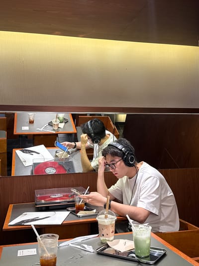 In a room with wood-paneled walls, a person wearing headphones and glasses is shown from the waist up. They are leaning forward, with their right hand extended towards a table. The table has several objects on it, including a red vinyl record in a clear plastic case, a record player, a phone, and two plastic cups with straws. One cup contains a green beverage, and the other contains a dark liquid. A white napkin with text is also on the table, next to a black watch on the person's wrist. The scene appears to be indoors, possibly a cafe or a personal space, and the lighting suggests it could be daytime. The person's posture and focus imply they are engaged in an activity, possibly related to music or work. The reflection in the mirror shows another person with headphones and glasses, facing the viewer, with a white t-shirt and black-rimmed glasses. The reflection also shows more of the room's interior, including wooden furniture and a mirror. The overall impression is one of concentration and engagement with the objects on the table.