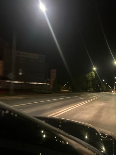 This is a night-time, motion-blurred photograph taken from inside a vehicle looking forward and slightly to the right. The viewpoint suggests the vehicle is moving along a multi-lane road. Streetlights, some with bright lens flares, illuminate the scene. To the left, a dark, multi-story building with some illuminated windows is visible. Trees line the right side of the road, and further down, a series of streetlights and lights from other vehicles are apparent. The road surface has visible white lane markings. The foreground is dominated by the dark, reflective surface of the vehicle's hood and windshield. The overall impression is of a journey through a moderately lit urban or suburban environment at night in Siedlce, Poland.