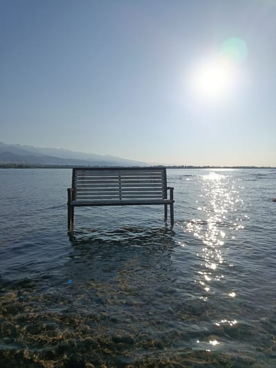 A wooden bench is submerged in shallow, clear water at the edge of a body of water, possibly a lake or sea, with mountains in the distance. The sun is shining brightly, creating a glare on the water's surface. The bench is a light color, with a slatted seat and backrest, supported by legs that disappear into the water. The water is calm and transparent, allowing the rocky bottom to be visible. The sky is a clear blue. The scene evokes a sense of tranquility and natural beauty. The location is identified as Kurskoe, Kyrgyzstan.