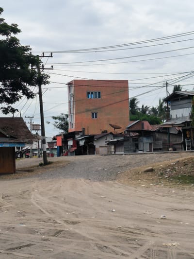 The image captures a daytime street scene in Bateiiliek, Indonesia, under an overcast sky. In the center, a four-story building with a peach or orange-brown facade and blue-tinted windows stands prominently. Several air conditioning units are visible on its exterior. Surrounding this central building are numerous smaller, older structures, likely houses or shops, characterized by their varied construction materials including wood, sheet metal, and corrugated iron roofs, many showing signs of wear. A satellite dish is mounted on the roof of one of these smaller buildings to the left of the central structure.
The foreground consists of a wide, unpaved dirt road or open area, deeply rutted with numerous tire tracks and strewn with scattered litter, including paper and plastic debris. The ground slopes slightly upwards to the right.
To the left, a large, leafy green tree partially obstructs a wooden building with a rusty corrugated metal roof. A tall power pole with multiple insulators and a dense network of overhead electrical wires dominates the left and central mid-ground, extending across the frame. Another power pole is visible further down the road. In the mid-left, near some of the smaller buildings, a person is seen riding a red motorcycle.
Visible text on a sign near the motorcycle reads "KINI LEBIH CEPAT" (Now Faster) and "MK CEL" and "Home XL Laga", indicating a commercial establishment, likely a mobile phone or internet service provider. Palm trees are visible in the background, contributing to the tropical setting. The scene depicts a blend of more modern and traditional structures, indicative of a semi-urban or rural area.
