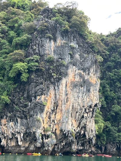 In Krasom, Thailand, a vertical rock face covered in lush green vegetation rises from the water. The rock face is gray and striated with streaks of orange and white. Several small kayaks are visible on the emerald green water in the foreground. These kayaks appear to be carrying people, suggesting a recreational activity such as kayaking or sightseeing. The dense green foliage on the cliffs indicates a tropical environment.