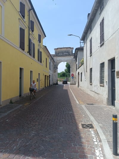 In Casalmaggiore, Italy, a cobblestone street is lined with buildings. On the left, a yellow building with dark-shuttered windows features a woman cycling with a basket on her handlebars. On the right, a pale building has dark-shuttered windows and a dark door. In the background, an archway frames a view of trees and a road with a stop sign and a black car. The sky is clear and blue.