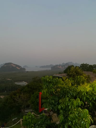 The scene shows an elevated view of Khlong Khian, Thailand, likely taken at dusk or dawn. The sky is overcast and grey. The foreground features dense green foliage and some buildings. In the distance, islands or land masses are visible amidst the water, partially obscured by the low light. A red object is seen among the trees in the near foreground.