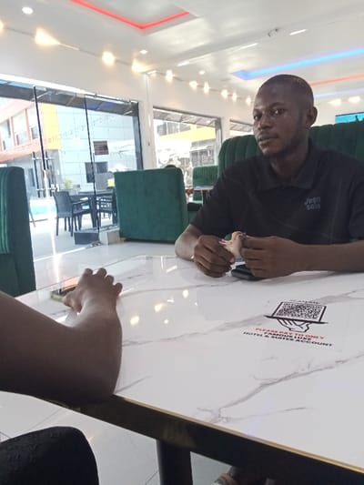The image shows two men sitting at a table inside a restaurant located at coordinates 5.471, 5.818 in Ujevwu, Nigeria. The man on the right is wearing a black polo shirt with the words "Jean Selia" printed on it. He is holding a cell phone in his left hand and has his right hand resting on the table. The man on the left is wearing a black shirt and has both arms on the table. The table is white with a black border and has a sticker that says "SCAN ME PLEASE PAY THROUGH ONLY FAMOUS LINK HOTEL & SUITES ACCOUNT". The background shows a large window with a view of a swimming pool and an orange building. There are green chairs around the table and a white ceiling with fluorescent lights.