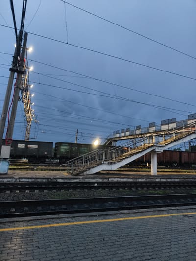 The image depicts a train station in Magdagachi, Russia, at dusk. In the foreground, a platform paved with grey interlocking blocks features a yellow safety line. Train tracks run parallel to the platform. A pedestrian overpass with concrete stairs and metal railings spans the tracks. Several freight cars are visible on the tracks, some in motion, blurred by the camera. Overhead power lines crisscross the sky. A utility pole with illuminated lamps stands to the left. The sky is overcast and dark blue. No people are visible.