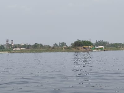 The image displays a wide horizontal view across a body of water, likely a river, in Narayanganj, Bangladesh. The lower half of the frame is dominated by the calm, greyish-blue water surface, exhibiting small ripples. The midground features the distant shoreline, presenting a mix of natural and industrial elements under an overcast, light grey sky.

On the left side of the visible shoreline, two prominent, tall, grey cylindrical structures with dark rectangular patterns are discernible, indicative of industrial facilities such as chimneys or cooling towers. Surrounding these structures are various smaller buildings and a line of green trees and sparse vegetation.

Towards the right side of the shoreline, a green and white-colored floating structure, possibly a moored barge or pontoon, is positioned near the bank. This structure features several yellow components on its deck. Slightly to its left, on the vegetated bank, a yellow tripod, consistent with a surveying instrument, is visible. The shoreline between these focal points consists of a low bank covered with green and brown vegetation, with scattered debris visible along the water's edge. No individuals are present or discernable within the captured scene. The upper portion of the image is occupied by a uniformly light grey sky.