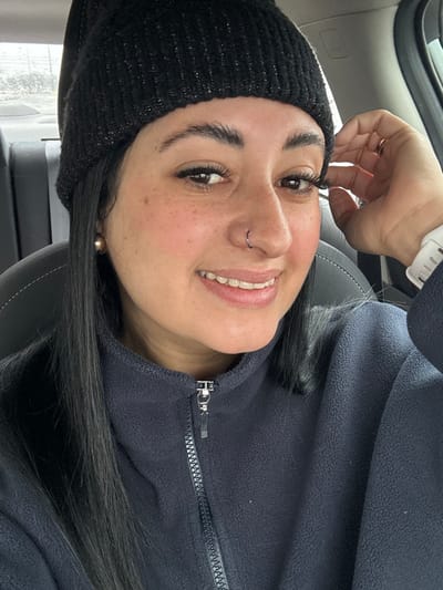 A smiling woman, wearing a black beanie and dark fleece jacket, is indoors. She is fair-skinned with dark hair and eyebrows, wearing a nose ring. The background features a car interior, with a grey leather seat. She is wearing a white smartwatch.