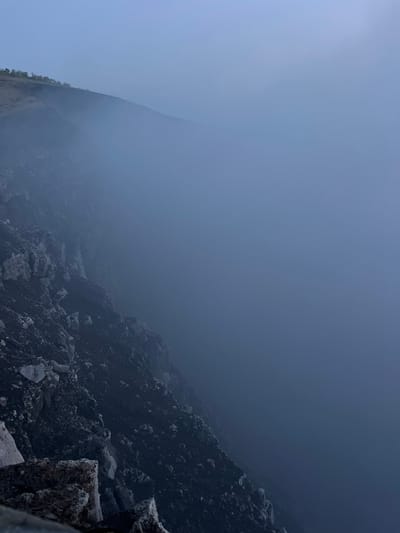 The image depicts a rugged, rocky mountainside partially obscured by a thick layer of fog or mist. The terrain on the left is dark and uneven, with visible rocks and dark soil. A sparse line of trees can be seen along the ridge in the upper left portion of the image. The rest of the scene is dominated by a hazy, blue-gray atmosphere, suggesting low visibility and a cool, possibly damp environment. The composition gives a sense of depth and a vast, open space obscured by the fog. There are no people or specific man-made objects visible, indicating a natural, possibly remote, landscape. The location is identified as being in the city of Nindirí, Nicaragua, though the image itself doesn't provide explicit visual cues to confirm this specific geographical context.