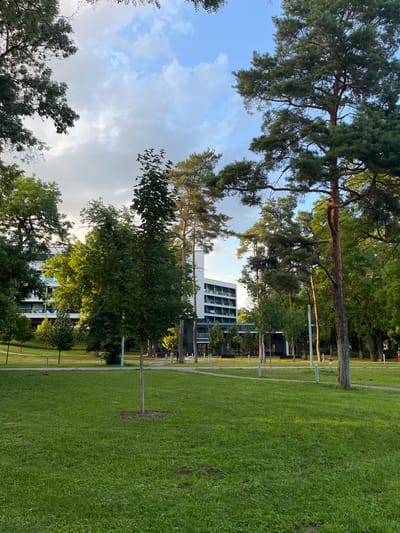 The image depicts a green park area in front of a modern white building with dark accents. The building has multiple floors with balconies and visible window panes. Several trees of varying heights, including tall pine trees and deciduous trees with full green foliage, are scattered throughout the park. The ground is covered in lush green grass, with some bare patches visible. A paved pathway winds through the park, and several light poles are present. In the background, two figures are walking on the pathway. The sky above is a mix of blue sky and white, gray clouds, suggesting either a partly cloudy or overcast day. The scene appears to be a well-maintained urban park or university campus, possibly located in Nyíregyháza, Hungary, given the context provided.