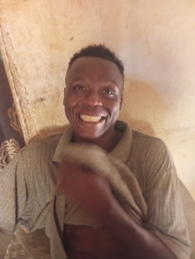 A smiling African man with short hair wears a collared shirt in front of a beige wall. His teeth are prominent in his bright smile. A partially visible patterned curtain is to the left. He holds a blurred white object in his hand, possibly a cloth. The image is taken indoors and appears to be somewhat informal.