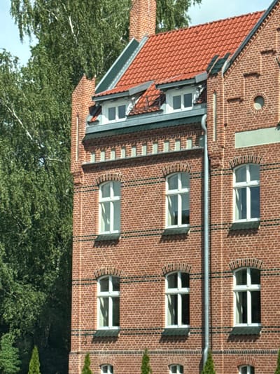 The image shows a close-up, eye-level view of a brick building with a red tiled roof and several windows. The building is made of red bricks with dark horizontal mortar lines. There are dormer windows with white frames on the roof. The main windows are also white-framed and appear to be double-paned. Some of the windows have arched tops. A metal downspout runs vertically down the side of the building. To the left of the building is a large tree with green foliage and white bark. The sky is visible and appears to be partly cloudy. The building is located in Šilutė, Lithuania.