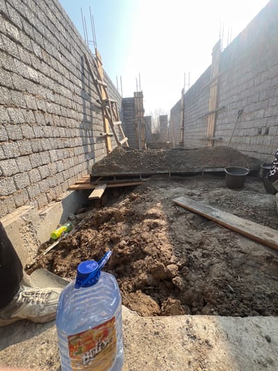 The image depicts an outdoor construction site in Дербісек ауылдық округі, Kazakhstan, under clear, bright daylight. The scene is dominated by two parallel, unfinished walls constructed from grey concrete blocks, extending into the background and forming a corridor. Vertical rebar protrudes from the tops of the walls at various points. A wooden ladder leans against the left wall. Wooden formwork is visible along the interior of both walls. In the central background, a large mound of dark brown soil and gravel sits on what appears to be a foundation or a recently filled area. Scattered wooden planks are present on the uneven dirt ground.
In the mid-right, a black bucket and the handle of a shovel are visible near the base of the right wall. A yellow tool, possibly a spirit level, lies on the ground near the base of the left wall. In the immediate foreground, a person's left foot, wearing a light-colored sneaker and dark trousers, is visible on a concrete surface. Next to the foot, a large clear plastic bottle with a blue cap and a multi-colored label (partially legible as "Бізнес") rests on the same surface. A small portion of a garment with a black and white pattern is visible in the bottom-right corner, near the black bucket. The ground throughout the site consists of uneven compacted dirt and loose soil.