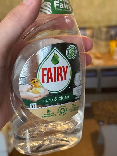 A hand holds a transparent plastic bottle of Fairy brand dish soap. The label reads "FAIRY" in large red letters with "pure & clean" underneath. A graphic shows a hand holding a small duckling. The label also includes green text and the words "95% биоразлагаемых ингредиентов" in Russian, indicating 95% biodegradable ingredients. The bottle is partially filled with clear liquid. The background is blurred and indistinct. There is no information in the image about the location of Chuprino, Russia.