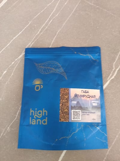 A blue pouch of Highland tea, labeled "GABA Izumrudnaya." Golden text reads "high land." The pouch has a tea leaf design and a cup with a sun logo in gold. A small window reveals the tea leaves inside. A label displays mountain imagery, a QR code, and "100G." The pouch is set against a grey, marbled surface.