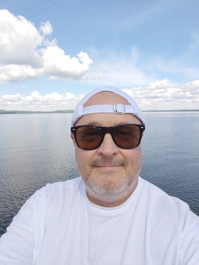 In this selfie, a man wearing a white baseball cap and sunglasses is captured from the chest up, standing outdoors. He has a light-colored beard and is dressed in a white t-shirt. Behind him, a large body of calm water stretches to the horizon, where a tree-lined landscape is visible under a bright sky dotted with white clouds. The man is looking directly at the camera, and his expression is neutral. The overall setting suggests a peaceful, outdoor environment, possibly a waterfront location. The image does not provide any specific clues to identify it as being in the city of Snezhinsk, Russia.