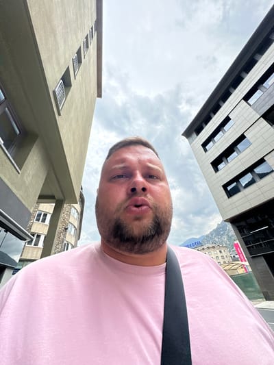 This image is a low-angle selfie, with a man's face prominently featured in the center-right, looking directly at the camera. He has light hair, a beard, and a mustache, with his lips slightly parted. He is wearing a light pink t-shirt and a dark strap, possibly from a bag, crosses his right shoulder.
The photograph is taken from a perspective looking upwards, showcasing an overcast sky with white and gray clouds occupying a significant portion of the frame. Several buildings surround the man. In the upper left quadrant, a modern building with dark and light paneling and rectangular windows is visible. To the lower left, an older-style building with a light-colored, possibly concrete, facade features small, symmetrically arranged windows with white railings. Behind the man's right shoulder, another building with a stone-textured or patterned facade and arched windows is discernible.
In the background, beyond the immediate buildings, mountainous terrain can be seen, hinting at an urban setting within a hilly or mountainous region. Partially visible and upside down on a distant building or sign, the text "NOVOTEL" and "MERCURE HOTEL" provides a significant clue to the location. These hotel names, coupled with the mountainous landscape, confirm the setting as Andorra la Vella, Andorra. The scene is illuminated by daylight, typical of a cloudy day. The man's expression is neutral and direct, suggesting a casual selfie.