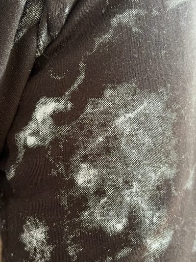 This is a close-up, high-angle, full-frame photograph of a dark brown fabric, likely clothing. The fabric has a textured weave, with visible knitting or threading. Scattered across the fabric are irregular, splotchy patterns of white or light gray material. These patterns appear to be printed or applied onto the fabric and have a slightly metallic or reflective sheen. Some areas show a higher concentration of the white material, forming denser patches, while others are more sparse and dispersed, resembling a marbling or tie-dye effect. The lighting suggests a single light source from the upper right, casting subtle shadows that highlight the texture of the fabric and the raised nature of some of the white patterns. The image does not depict any recognizable landmarks or features of the city of Hanniv, Ukraine, and focuses solely on the material's surface.