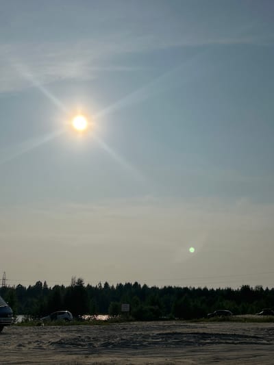 The image depicts an outdoor scene on a sunny day, likely during the late afternoon or early evening due to the sun's position and the light quality. The sky is a pale blue with some wispy clouds. The sun is bright and positioned in the left half of the frame, casting a strong glare.
The foreground of the image shows a rough, sandy or dirt surface with tire tracks, suggesting it might be a dirt road or a parking area. To the right, a dense line of dark green trees forms a treeline, with some lighter foliage interspersed. Beyond the trees, there are hints of what could be a body of water reflecting the sunlight, creating a shimmering effect. Several dark-colored cars are parked along the edge of the treeline, and one white car is partially visible. There is also a tall, lattice-like electrical pylon in the background.
The overall atmosphere is one of a clear, bright day in a somewhat rural or undeveloped area, possibly on the outskirts of Noyabrsk, Russia, given the provided context. The bright sunlight and the presence of trees and a dirt road are typical of such a location. There is no clear activity or event taking place; it appears to be a still moment captured outdoors. No legible text is visible in the image.