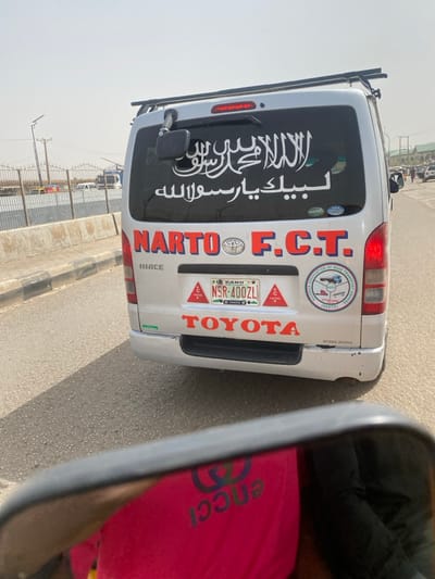 The image depicts the rear of a silver Toyota Hiace passenger van (mini-bus) on a paved road in Gobirawa, Nigeria, under bright daylight conditions. The van features a roof rack and a large white Arabic calligraphy decal on its black rear window, reading "لَا إِلَٰهَ إِلَّا ٱللَّٰهُ مُحَمَّدٌ رَسُولُ ٱللَّٰهِ" (There is no god but Allah, Muhammad is the messenger of Allah) and "لبيك يا رسول الله" (Here I am, O Messenger of Allah). Below the window, "NARTO F.C.T." is emblazoned in red letters, with a circular emblem to the right indicating the "NATIONAL ASSOCIATION OF ROAD TRANSPORT WORKERS (NARTO) FCT BRANCH". The vehicle's license plate, located centrally, reads "KANO NSR-400ZL". Three red triangular "CAUTION" stickers are affixed around the license plate. The word "TOYOTA" is prominent in red below the license plate. The lower rear bumper shows minor scuff marks. To the left of the van, a concrete road barrier is visible, and the background reveals a wire fence, utility poles, and distant structures, indicating an urban environment. The photograph is taken from a low perspective, with a partial reflection in a mirror at the bottom of the frame showing a person wearing a pink garment with the reversed "GUCCI" logo. No specific individuals are discernible inside or immediately outside the van.