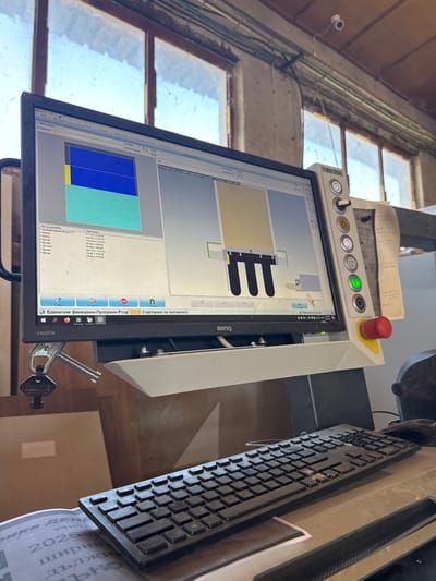The image shows a close-up of a computer monitor and keyboard in what appears to be an industrial or workshop setting. The monitor displays a technical interface with a diagram of what looks like a manufactured part, along with lists of materials and dimensions. The brand "BIESSE" is visible on the side of the equipment. A set of keys hangs from a clip below the monitor. The background suggests a room with large windows and exposed pipes, hinting at an older building. The presence of a keyboard and monitor indicates an active workspace. The overall scene is one of a functional, operational environment. The user has provided information that this location is in the city of Belene, Bulgaria, however, there are no visual cues in the image that corroborate this specific location.