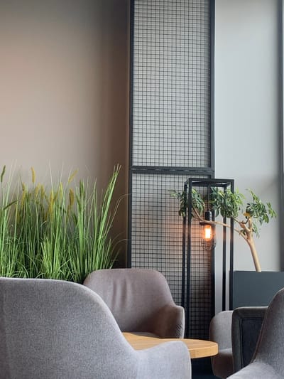 The image shows a modern interior design of a café or restaurant in Pyrzowice, Poland, located at latitude: 50.471, longitude: 19.072. The scene is captured from a low angle, showcasing a large window on the right, with a white wall behind it. To the left of the window, a black metal mesh screen is positioned against a light brown wall. Two grey upholstered chairs are facing each other, with a wooden table in between. A black metal lamp stand is positioned between the chair on the right and the mesh screen. The lamp has a large, clear lightbulb emitting a warm yellow glow. A potted plant with small green leaves is situated on the right of the lamp, behind the chair. A large plant with long, thin green blades is placed in a pot on the left side of the image, against the light brown wall. The interior has a minimalist, modern aesthetic, with clean lines and simple colors. There are no people present in the scene.