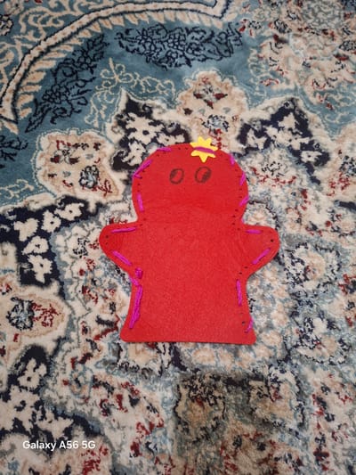 A red felt doll, with pink stitching along its edges and a yellow star on its head, lies on a patterned blue and white rug. The doll has two round, brown eyes and a star-shaped yellow cutout on its head. The rug has an intricate floral and paisley design. The bottom left corner of the image has a "Galaxy A56 5G" watermark. This image is not located in the city of Greater London, United Kingdom.