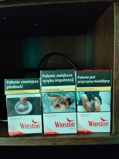 Three packs of Winston cigarettes are arranged side-by-side on a dark wooden shelf. The packs have graphic warnings in Polish. The leftmost pack has a warning about reduced fertility and an image of a hand emptying an ashtray. The middle pack warns about increased risk of impotence and shows a graphic of a person lying in bed. The rightmost pack states that smoking causes gangrene and displays an image of a gangrenous foot. The background is dark, with some subtle teal lighting, and a dark cable is visible behind the cigarette packs. The scene does not provide any specific information that would allow for localization in Płock, Poland.