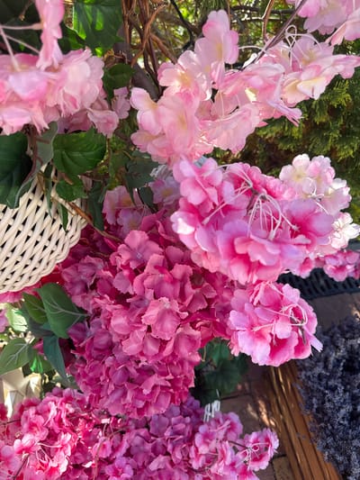 This is a close-up, eye-level, outdoor shot of artificial flowers, predominantly in shades of pink, with some green foliage visible. The flowers appear to be a mix of hydrangeas and possibly cherry blossoms, densely packed together. In the upper left quadrant, a portion of a white woven basket is visible. To the right, a dark green coniferous plant is out of focus. In the lower right, a bundle of dried lavender, a dark purple, is present. The background suggests a brick or stone surface with wooden elements. The lighting indicates it is daytime, possibly late afternoon due to the warm tones and shadows. The location is identified as Berehove, Ukraine. There are no people or animals in the image.