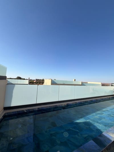 The image depicts a rooftop infinity pool located in Tassoultante, Morocco, under a clear blue sky. The pool occupies the lower half of the frame, featuring dark blue and slate-patterned tiles on its bottom and sides, with water reflecting the sky. A white, circular pool drain is visible on the pool floor in the lower right foreground. Along the far edge of the pool, a modern railing composed of light blue, translucent glass panels with black trim runs horizontally across the midground. Beyond the railing, the cityscape of Tassoultante is visible, characterized by a mix of light-colored, low-rise buildings and scattered green vegetation. The upper two-thirds of the image is dominated by a cloudless, bright blue sky, indicating clear daylight conditions. No individuals are present in the scene, and no actions or interactions are depicted.