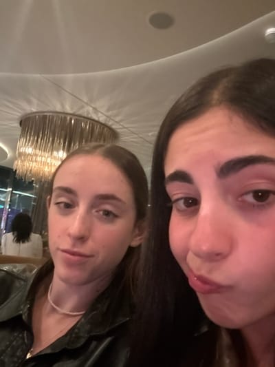 Two young adult females are featured in a close-up indoor photograph. The subject on the left, with light brown hair pulled back, wears a dark jacket and a light-colored beaded necklace, looking towards the camera with a neutral expression. The subject on the right, partially visible on the right side of the frame, has dark hair and is making a puckering gesture with her lips. In the blurred background, a third individual is seen from behind, wearing a white top, seated in what appears to be a dining or lounge setting. A large, multi-tiered chandelier with numerous vertical, reflective elements hangs from a curved white ceiling, casting distinct light patterns. Recessed ceiling lights or speakers are also visible. The environment suggests an upscale restaurant or lounge. The image provides no discernible information to confirm its location in Rum Cay, Bahamas.