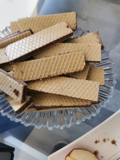 A close-up, slightly angled shot shows a pile of chocolate-filled wafer cookies on a clear, fluted glass plate. The cookies are rectangular with a textured, waffle-like surface and have a dark chocolate filling visible between the layers. Some of the cookies are broken. The plate is placed on a reflective surface that appears to be a table. In the background, a blurry, light blue surface is visible. In the lower right corner of the image, a portion of a white plate with crumbs and a slice of apple is visible. The image is well-lit, highlighting the details of the cookies and the glass plate. The setting does not provide any specific clues that would allow for the determination of a location within the city of Bourg-lès-Valence, France.