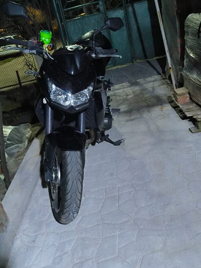 A black motorcycle is parked facing forward on a light-grey, polygonal-patterned paved surface in Dolistovo, Bulgaria. The motorcycle features dual illuminated headlights, a visible radiator, black handlebars with rearview mirrors, and a distinct neon-green cylindrical fluid reservoir mounted on the left handlebar. The front tire displays a noticeable tread pattern. In the left background, a section of metal mesh fencing is partially visible. In the right background, a dark metal gate is present, and further to the right, a stack of items, possibly wooden pallets, wrapped in clear plastic, rests on the ground, supported in part by a red brick. The scene is illuminated by artificial light.