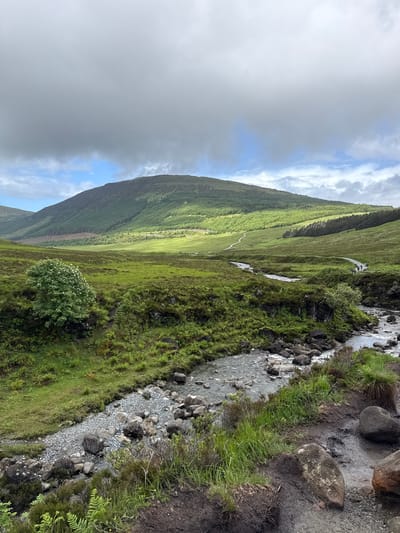 The image depicts a scenic landscape on the Isle of Skye, United Kingdom. A winding river flows through a valley, with rocky banks and lush green vegetation. In the background, a large, verdant hill rises, partially covered by a dense forest of pine trees. The sky above is a mix of dramatic clouds and patches of blue, with sunlight casting shadows and highlighting parts of the landscape. A narrow path can be seen winding up the hill, with a few distant figures walking on it. The foreground features more rough terrain with rocks and dense green undergrowth.