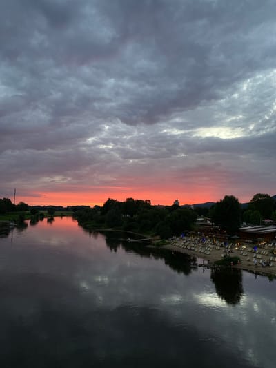 This photograph depicts a twilight scene over the Weser River in Rinteln, Germany. The sky is filled with dramatic, heavy clouds, tinged with hues of gray and muted pinks and oranges from the setting sun. A vibrant band of red-orange stretches across the horizon, reflecting vividly on the calm surface of the river.
On the right bank of the river, a sandy beach is illuminated with numerous lounge chairs and umbrellas, suggesting a recreational area. A cluster of buildings, possibly restaurants or beach clubs, with lights on, are visible along the shoreline. Several individuals are scattered across the beach, some sitting in chairs, others standing or walking.
The left bank of the river is lined with dense green trees and foliage, with a solitary tall structure, likely a cell tower, standing out against the dusky sky. The river itself is dark and reflective, mirroring the sky and the trees. The overall impression is one of a serene, late-evening atmosphere at a popular riverside location.