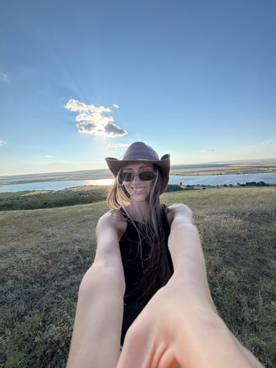 A selfie is taken by a woman wearing a brown cowboy hat and sunglasses, a black tank top, and a wide smile. She is standing on a grassy hill overlooking a wide river or lake with distant, sparsely vegetated shores. The sky is blue with scattered clouds and the sun is shining brightly. The woman's arms are extended forward, holding the camera. The environment appears to be rural, with dry grass covering the foreground and middle ground. The lighting suggests it is late afternoon or early evening. The location is the city of Gornyi Balyklei, Russia.