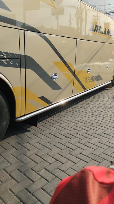 The image shows a side view of a beige bus with black and yellow graphic accents, parked on a herringbone brick paved surface. The bus has several doors with silver handles and a chrome running board along its lower side. The text "AIRLANDER" is visible on the upper right side of the bus. The environment appears to be outdoors, possibly a street or parking area, with reflections of overhead wires and trees visible on the bus's surface. A portion of a red object, possibly a piece of clothing or bag, is in the foreground, out of focus. The scene is captured in daylight.