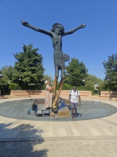 In the city of Međugorje, Bosnia and Herzegovina, a large, dark bronze statue of a crucified Jesus stands prominently in an open plaza. The statue is elevated on a golden base and is surrounded by a circular tiled area. Two individuals are present near the base of the statue. On the left, an elderly woman wearing a patterned dress and a hat is standing on a small step, appearing to interact with the statue. Beside her is a blue walker with a black bag attached. To the right of the statue, a Black man in shorts and a white polo shirt with a backpack is standing and looking towards the statue. The plaza is bordered by low walls with benches and is surrounded by trees and a clear blue sky. The sun is shining, casting shadows on the ground.