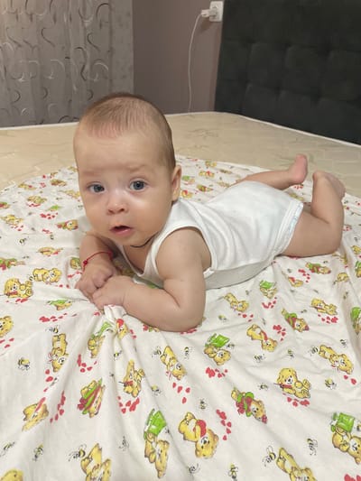 The media file shows a baby lying on its stomach on a white crib sheet with a pattern of teddy bears and bees. The baby is wearing a white onesie. The baby has light brown hair and blue eyes and appears to be looking at the camera with a curious expression. Its mouth is slightly open. A red string bracelet is visible on its left wrist. The background is out of focus but appears to be a wall and possibly part of a bed. The lighting suggests it is daytime indoors. The scene is casual and domestic, likely in a home setting. There is no visible text in the image. The location context is Fălești, Moldova.