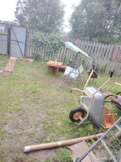 This media file depicts an outdoor scene in what appears to be a backyard in Gorshki, Russia. The setting is a somewhat overgrown yard with patches of mud, suggesting recent rain or damp conditions. The sky is overcast, indicating a cloudy day, possibly morning or late afternoon.
In the foreground, a wheelbarrow is prominently placed, tilted on its side. It is a light-colored metal wheelbarrow with a black tire and an orange rim. Next to the wheelbarrow, there is a brown cardboard tube lying on the ground and parts of a metal ladder are also visible. Further back, a wooden table with a reddish-pink cushion is visible, with a glass or cup on top.
To the left, there are two gates, one a large corrugated metal gate and another section of fence with metal bars. Beyond the fence, tall green trees form a dense backdrop. In the middle ground, a garden swing set with a green canopy and a striped cushion is visible. A small, dark barbecue grill on legs is also present behind the swing. There are some plants growing along the weathered wooden fence that runs horizontally across the middle of the frame. A piece of patterned fabric, possibly a rug or blanket, is draped over the fence on the right side.
The overall impression is of a somewhat cluttered, functional outdoor space, possibly belonging to a dacha or a rural residence. No people are visible in the image, and the scene appears to be static, with no specific activity taking place. The mood is muted, reflecting the overcast weather. There is a visible label on the side of the wheelbarrow, but the text is illegible.