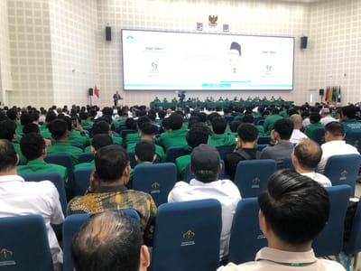In the city of Reuleut Timu, Indonesia, an indoor auditorium is filled with attendees seated in rows of blue chairs. A large screen at the front displays text and an image of a man in traditional attire, with the text "KAPAL TERBUKA" and "57 MIMARA SEJARAH". A speaker stands at a podium on a stage to the left of the screen. The audience consists primarily of young men, many wearing green shirts, facing the stage. The auditorium walls are white and textured, and the lighting suggests an artificial illumination. The overall scene depicts an organized event or conference.