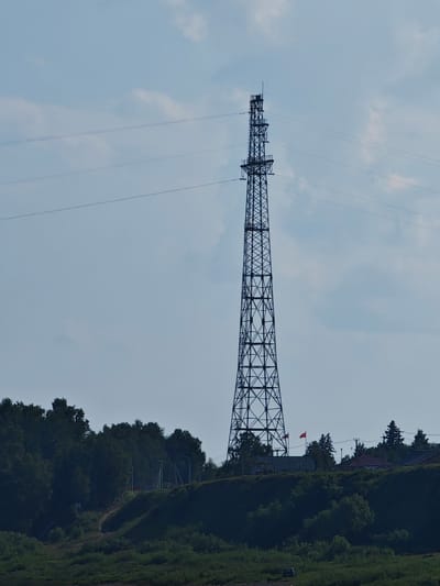 This is a vertical, eye-level, full shot of a tall, dark metal lattice power pylon set against a pale blue sky with scattered white clouds. The pylon is the dominant feature in the image, located on the right side of the frame. Three thin power lines extend horizontally from the top of the pylon and disappear out of frame to the left. Below the pylon, on a green, tree-covered hillside, are several small buildings and two flags, one red and one appearing to be the Russian flag. A dark, dense line of trees runs along the crest of the hill. In the foreground, a body of water with a dark green grassy bank is visible. The scene appears to be in Верхнепашино, Russia.